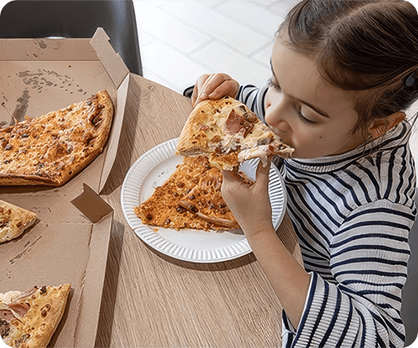 A girl eating pizza at a table with two boxes of pizza.
